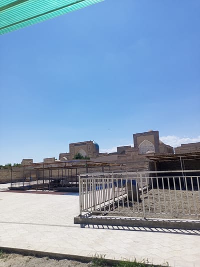 The image captures an outdoor scene in the city of Shar-Bakr, Uzbekistan, under a clear blue sky. In the foreground, a paved area with large, light-colored tiles extends across the lower portion of the frame. A concrete curb separates the tiled area from a strip of sandy ground with sparse grass. A white metal fence runs horizontally across the middle ground, enclosing an area with a sandy floor and several structures that appear to be animal enclosures or stalls with open sides and roofs supported by metal poles. Some large blue barrels are visible within these enclosures.

In the background, several historically significant buildings are visible. These structures are constructed of brick and feature arched doorways and windows. One prominent building has a dome topped with a blue tile accent. Another building features intricate tilework around a large archway. The architecture suggests a religious or historical complex. The overall environment appears to be sunny and dry, consistent with a desert climate. There are no people visible in the image.