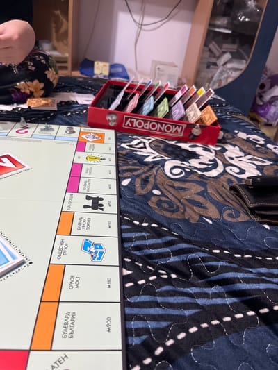 A Monopoly game is in progress, with the board displaying Bulgarian street names such as Bulevard Evlogi Georgiev and Bulevard Hristo Botev. A red Monopoly money holder with stacks of colorful paper currency stands beside the board. The game is being played on a quilted surface with a brown, blue, and white patterned design. Part of a person's arm and a floral-patterned sleeve are visible at the bottom left. In the background, there is a shelf with miscellaneous items.