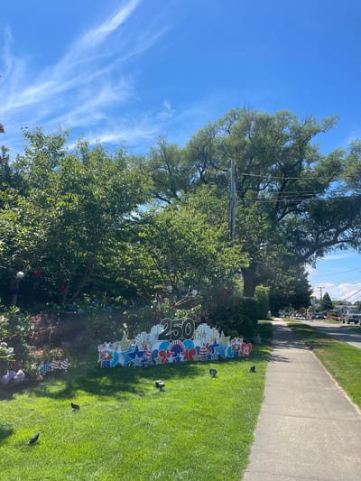 This image captures a patriotic display in a suburban setting, likely Edgartown, USA, on a sunny day. The primary subject is a yard decoration spelling out "250" adorned with stars, stripes, and fireworks motifs in red, white, and blue, celebrating a significant anniversary. Smaller patriotic-themed signs, including American flags and stars, are scattered around the lawn, accentuating the festive theme.

The scene unfolds on a well-maintained green lawn bordering a paved walkway. Lush green trees provide a backdrop, with a large tree prominently featured on the left, casting shadows on the grass. The sky is a vibrant blue with wispy white clouds, suggesting fair weather. In the distance, a street lined with houses and parked cars hints at a residential neighborhood.

The overall mood is celebratory and patriotic. There are no visible people in the immediate foreground, but the decorations suggest a community or individual is marking a special occasion. The time of day appears to be late morning or early afternoon, given the bright sunlight and clear sky. The decorations, particularly the "250" sign, indicate a celebration of the 250th anniversary of something significant, possibly a town or a nation.