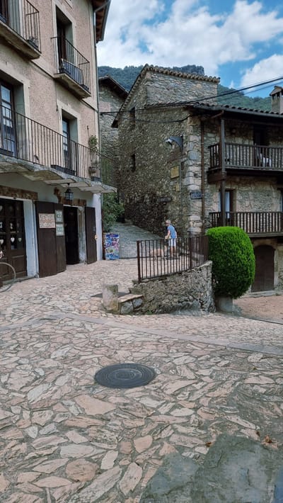 This image depicts a street scene in the city of Camprodon, Spain. The foreground is paved with irregular stone slabs and features a circular metal manhole cover. A person in a blue shirt and white shorts is walking a dog up a sloped stone pathway in the midground. They are on the right side of a dark metal railing. To the left of the pathway are buildings constructed with stone and stucco. One building in the left midground has a sign that reads "VENDA EMBOTITS PRODUCTES DE LA CERDANYA". Further back, more stone buildings are visible, with a mountain landscape in the background under a cloudy sky. The overall impression is of a historic village street.