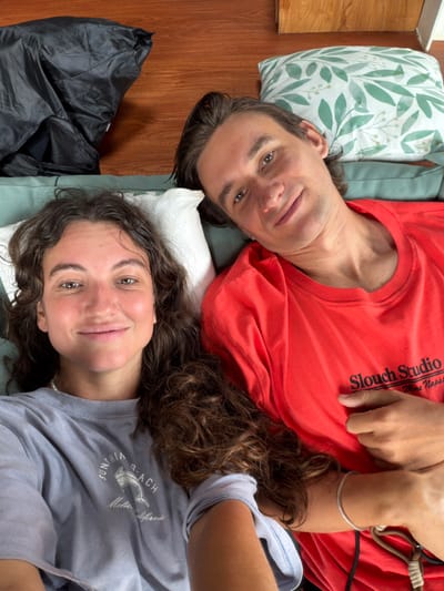 Two individuals lie on their backs, resting their heads on pillows. One wears a red shirt with "Slouch Studio Wine Napa" printed on it. The other person is in a grey shirt, and some text printed that is not readable. They both look up at the camera. A dark bag sits on the floor next to them. There are some bedsheets and the pillow has a green pattern. The floor has some wood.