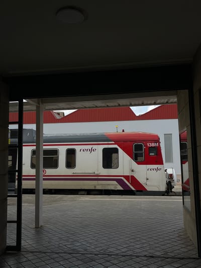 The image displays a Renfe commuter train at a railway station in Buñol, Spain, viewed from within a darkened interior space. The foreground consists of a dark ceiling with a circular light fixture, dark overhead framing, and a grey tiled floor with a grid pattern. A white vertical column is visible on the left side of the frame.
The central subject is a Renfe train car, predominantly white with red and purple diagonal accents. The word "renfe" is visible on its side in purple lettering, and the identification "138M" is clearly marked on the front-facing section of the car. The train features multiple rectangular windows along its side and a dark grey roof with a small yellow antenna-like object. It rests alongside a grey tiled platform, which transitions from the interior flooring.
In the background, beyond the train, are several white buildings topped with distinct red corrugated roofs, exhibiting a sawtooth profile. A narrow, vertical rectangular window is visible on one of the white structures. A small segment of pale blue sky with white clouds is visible between the rooflines. No individuals are present in the scene, and no discernible actions or interactions are occurring.
