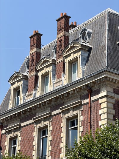 This image, taken in Courbevoie, France, shows a close-up, angled view of the upper facade of a brick building with stone architectural details. The building has a dark gray, tiled roof and multiple chimneys made of red brick with decorative stone caps. Several windows are visible, some with white frames and blinds, others open. The building's facade is adorned with elaborate stone moldings and cornices, creating a classical architectural style. A section of a green leafy tree is visible on the right side of the image, suggesting an outdoor setting. The sky is a clear, bright blue, indicating it is daytime, likely sunny. There is no activity or event depicted, and no text is visible. The orientation of the image is rotated, making it appear sideways.