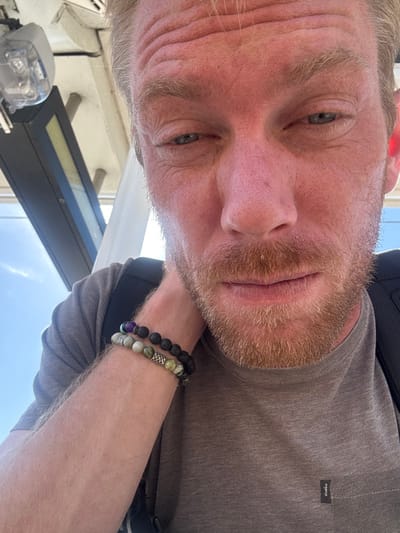 An adult male with a fair complexion, reddish-blond hair, and a short beard is captured in a close-up, slightly low-angle photograph. He wears a grey short-sleeved t-shirt, visible over his torso, and a dark backpack is partially discernible over his right shoulder. On his left wrist, two beaded bracelets are worn: one composed primarily of dark, possibly lava, beads with a single prominent purple bead and a silver-colored cylindrical spacer bead featuring an etched pattern; the second bracelet consists of mottled green and grey beads with a similar silver-colored cylindrical spacer. The subject's eyes are directed downwards, and his facial expression is neutral.
The background indicates an outdoor or semi-outdoor public space under an overhead structure. Visible architectural elements include a white overhead canopy, two grey vertical support poles, a dark rectangular sign or fixture, and a clear plastic-encased device, potentially a light fixture or security camera, mounted on the canopy. A clear blue sky is visible in the upper left background. These structural details, particularly the canopy and signage, are characteristic of the Long Beach Transit light rail stations, specifically the Downtown Long Beach Station or adjacent stations along the transit corridor in Long Beach, California, United States of America.