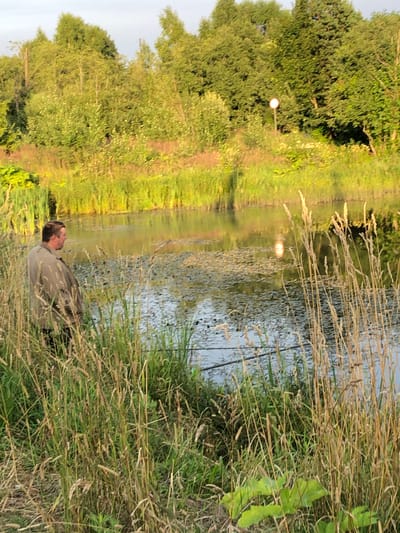 The image depicts a tranquil natural scene near the city of Makeikha, Russia, likely during the late afternoon or early evening given the warm lighting. In the foreground, a man, appearing to be in his late 30s or 40s, stands partially obscured by tall, dry grasses, facing away from the camera and towards a body of water. He is wearing a light brown jacket. The water is calm, reflecting the overcast sky and surrounding lush green trees. The shoreline is densely vegetated with reeds and other wild plants. In the background, a dense forest borders the water. A white circular sign on a pole is visible in the middle distance, surrounded by greenery. The overall impression is one of peaceful, natural beauty, perhaps a fishing spot.