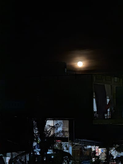 This image depicts a night scene in Lý Sơn Special Administrative Region, Vietnam. A bright full moon is prominently featured against a dark sky, with a hazy glow surrounding it. The moon illuminates the surrounding area, casting light on a multi-story building and some vegetation.
The building appears to be residential or commercial, with windows and balconies visible. Some windows are lit from within, suggesting activity inside. A metallic structure, possibly scaffolding or a utility pole, is also visible near the building. To the right of the building, there are signs of an urban environment with what looks like a marketplace or shop area, with lights and items for sale. A palm tree is also visible, its fronds silhouetted against the night sky.
The overall atmosphere is quiet and peaceful, with the moon being the dominant subject. The time of day is clearly night, as indicated by the darkness and the presence of the moon. There are no people actively visible in the frame, but the lit windows suggest human presence. The weather appears clear, allowing for a bright view of the moon. The scene is characteristic of a tropical or subtropical location due to the palm tree. No text is legible in the image.
