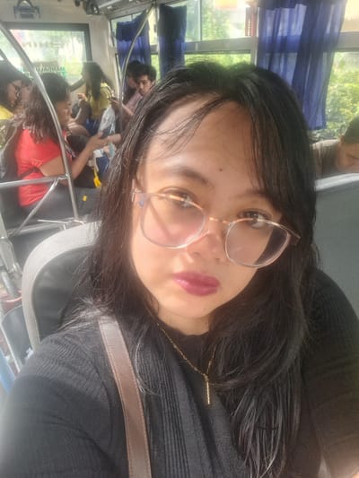 The media file shows a close-up, vertically oriented selfie of a person inside what appears to be a bus. The subject is a woman with long, dark hair, wearing glasses with light-colored frames and a black, ribbed long-sleeved shirt. She has on lipstick that is a deep pink or berry shade. A thin gold chain with a small bar pendant is visible around her neck. A brown strap, possibly from a purse, crosses her chest.

In the background, other people are visible, also appearing to be passengers on the bus. One person in the upper background is wearing a bright red shirt. The interior of the bus has blue seats and windows. Through the windows, greenery and trees can be seen, suggesting it is daytime and outdoors, possibly in a tropical or temperate climate. The overall lighting suggests it is a bright day. The reflection in the window shows more passengers and the bus interior. No discernible text is visible. The scene appears to be a casual moment captured while traveling, with the subject taking a selfie.