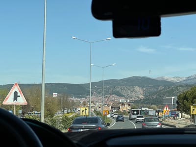 The image depicts a view from inside a car driving on a road in Menteşe, Turkey. The car is one of several in traffic on a multi-lane road. A gray sedan with license plate "48 ANG 962" is directly ahead. Road signs are visible on both sides of the road, including a triangular sign warning of a side road joining from the left and a sign indicating a speed limit of 30 km/h. A city with a mix of low-rise and taller buildings lies in the distance, backed by mountains. Overhead streetlights line the road. The sky is clear and blue. A small digital device is attached to the rearview mirror, displaying the number "10". A crane is working on construction of a tall building in the distance.