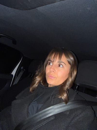 A young woman with brown hair and bangs glances at the camera, seated in a car. She wears a dark coat and a seatbelt is fastened across her chest. The car's interior is dark, with a black seat and ceiling. A white panel and a door handle are visible in the background. The image is angled, giving a tilted perspective.