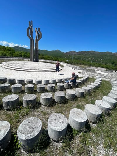 The image shows the Barutana Memorial Park in Montenegro. The park consists of a large circular concrete platform with a monument featuring three stylized abstract shapes extending upwards. A semicircle of concrete cylinders surrounds the platform, with grass growing between them. Two people are present: one stands near the monument and the other sits on one of the cylinders. In the background are mountains and a clear blue sky.
