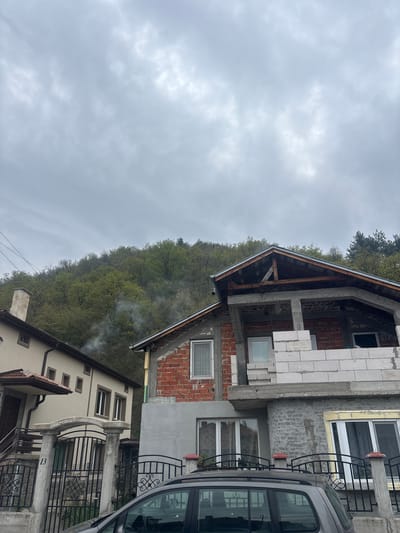 The image depicts a view in Bratsigovo, Bulgaria, under a cloudy sky. A building under construction dominates the scene, exhibiting a partially completed brick and cinder block facade with visible window openings. To the right of the construction, a partially visible silver vehicle is parked. Adjacent to the construction, another residential building with a wrought iron fence and balcony is present, displaying an address number "13." Smoke rises from an unseen source, drifting amongst the trees covering a hill that forms the backdrop. The vegetation indicates a deciduous forest in an early spring state.