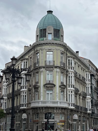 The image depicts the iconic building at Plaza de la Escandalera in Oviedo, Spain, known for its distinctive green dome and ornate architecture. The building, with its cream-colored facade and elaborate balconies, stands against a cloudy sky. A multi-armed lamppost with several lanterns dominates the left side of the frame, partially obscuring the building's facade. Several businesses are visible on the ground floor, including "LA CUPULA" and "ALIMERKA." A few pedestrians are present on the sidewalk in front of the shops, with traffic lights visible in the lower right corner. The architectural style is eclectic, featuring elements of Art Nouveau and neoclassical design.