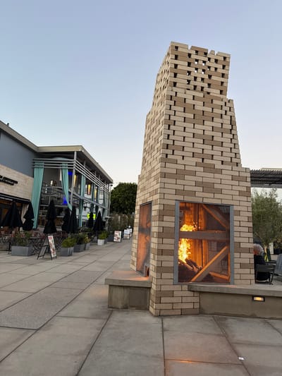 A tall, brick fireplace with an open fire is the central focus of this outdoor scene. The fireplace is constructed from light brown and tan bricks, with alternating patterns of openings near the top. A metal mesh covers the front opening where flames are visible. The fireplace sits on a concrete base, with a person with gray hair seated in a chair to the right.
To the left of the fireplace is a sidewalk paved with large concrete slabs. Further to the left, there is a building with large windows and teal curtains. Outdoor seating areas with tables and black umbrellas are set up in front of the building, suggesting a restaurant or cafe. Several signs are visible in front of the seating area.
The background shows more buildings and trees. The sky is a clear, pale blue, indicating it is either early morning or late evening. The overall impression is a pleasant, urban outdoor dining or gathering space. The location is identified as Manhattan Beach, United States of America.