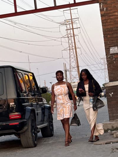 Two women stand on a sidewalk beside a black SUV. The woman closest to the camera wears a short, patterned dress and black heels, carrying a black tote bag. The second woman is behind her, wearing a long, patterned dress with a black jacket and carrying a shoulder bag and a water bottle. Utility poles and wires fill the sky in the background. A sign is partially visible on a building in the upper left corner. The setting appears to be an urban area.