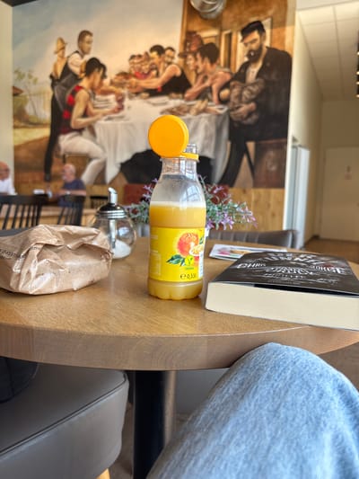 A person in blue jeans sits at a wooden table, partially obscuring a book and a bottle of orange juice. The book's cover displays the title "CHRIS DAME GEORG KNAUR THRILLER". The orange juice bottle has a yellow cap and a label featuring an orange. Also on the table is a small sugar dispenser and a brown paper bag. In the background, a mural depicts a scene with several people. The lighting suggests it is daytime.
