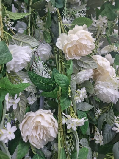 The media file is a close-up, high-angle shot of a floral arrangement. The subjects are artificial flowers and greenery. There are several large, creamy white peonies or roses, and smaller clusters of delicate white flowers that resemble jasmine or baby's breath. Lush green leaves, some with a slightly fuzzy texture, are interspersed throughout the arrangement. Thick, woven green stems are visible, forming a base or structure for the foliage. The lighting is even and appears to be artificial, suggesting the arrangement might be indoors or in a studio setting. There are no people, animals, or discernible objects other than the floral elements. The scene is static, with no activity taking place. The overall impression is one of artificial elegance and abundance. The location context of Wootton, United Kingdom, does not provide any visual cues within the image itself. No text is visible in the media file.