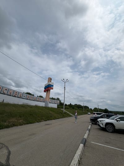The image depicts a street scene in Nikolaevka, Russia, under a cloudy sky. A large, elevated sign with Cyrillic lettering reads "МОСКВА-ВЛАДИВОСТОК," indicating the route connecting Moscow and Vladivostok. A tall pole with three horizontal colored panels, resembling the Russian flag, stands next to the sign. Several cars are parked on the right side of the road, including a white SUV in the foreground. A young woman in a white shirt and light-colored pants stands on the side of the road, facing away from the camera. Another person is visible near the parked cars. The road surface appears to be asphalt, and a grassy embankment with trees lines the left side of the road, leading up to the sign. Streetlights are visible in the background.