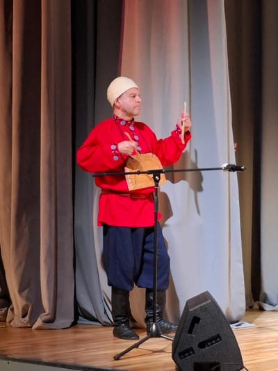 A man in traditional Russian attire stands on a wooden stage in front of a backdrop of curtains. He is wearing a red tunic with blue embroidered accents, a red belt, dark blue pants, black boots, and a white skullcap. He holds a wooden drum and a beater in his hands, appearing to be in the middle of a performance. A microphone stand is positioned in front of him, and a black speaker is visible in the foreground. The image was taken in Kungur, Russia.