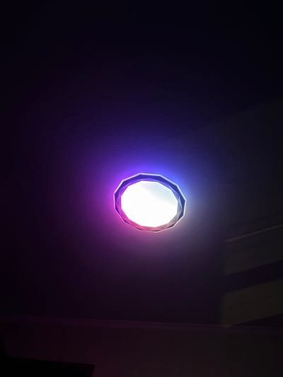 A ceiling light fixture is illuminated with purple and white light, casting shadows and reflections on a textured ceiling. The light fixture has a faceted, polygonal shape and appears to be recessed into the ceiling. The surrounding area is dark, with some lighter horizontal lines visible on the right side of the image, possibly indicating architectural features or reflections. The overall scene is dimly lit and lacks any visible people or specific environmental context that would allow for a precise location within Daugavpils, Latvia.