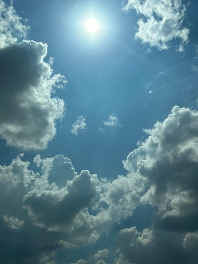 This media file depicts a view of the sky with the sun and clouds, taken from a low angle looking upwards. The sun is prominently featured on the left side of the image, emitting bright rays and lens flare. Various types of clouds are visible, ranging from bright, white, fluffy cumulus clouds to darker, more ominous storm clouds. The sky is a clear, vibrant blue. The scene suggests a day with dynamic weather conditions, possibly transitioning between sunny periods and potential rain. The time of day appears to be daytime, given the brightness of the sun. There are no people or objects other than the natural elements of the sky. The image is oriented such that the sun is in the lower left quadrant, and the clouds dominate the upper right and lower portions of the frame. The lighting and cloud formations create a sense of depth and scale, emphasizing the vastness of the sky. No text is visible in the image. Given the location context of Загорци, Bulgaria, this scene represents a typical skyscape that might be observed in that region.