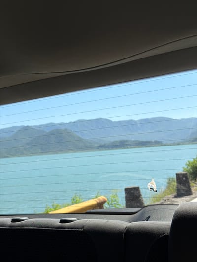 The image is a view from inside a vehicle, looking out through the rear window. The window has horizontal defroster lines. Outside, there is a body of water, possibly a lake, with mountains in the background. The water is a vibrant turquoise color, and the mountains are hazy and layered, indicating distance. There is some vegetation and what appears to be a yellow railing or barrier in the foreground. On the rear window, there is a small, light-colored object with dark markings, resembling a butterfly or a piece of debris. The interior of the car is visible at the bottom of the frame, showing the tops of dark-colored seats. The overall scene suggests a scenic drive through a natural landscape. The specific location of Kruševica, Montenegro, is not discernible from the visual elements within the image itself, as there are no identifying landmarks, signs, or distinctive geographical features that specifically point to this location.