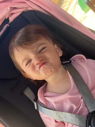 A close-up, high-angle shot shows a young child with brown hair and closed eyes making a funny face, with their cheeks puffed out and mouth pursed, as if holding their breath. The child is wearing a pink shirt and is secured by a gray stroller harness. The background is dark, with a pink fabric canopy visible above and a slightly blurred outdoor environment with yellow and green elements beyond. The scene takes place in a stroller, likely in a park or outdoor public area in the city of Elena, Bulgaria.