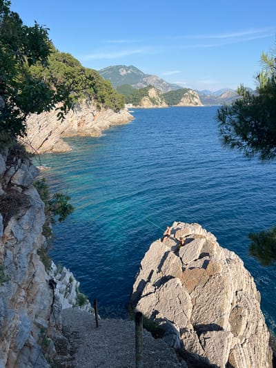 A high-angle, landscape-orientation photograph captures a rocky coastal area, possibly near Medinski Krš, Montenegro. The deep blue sea occupies a significant portion of the image. Rocky cliffs with sparse vegetation are seen on both sides. In the upper-right corner, two figures are seen sitting on a rocky outcrop. A thin, light-colored line appears to be attached to one of the figures, possibly a rope or wire. The sky is clear blue, and a distant mountain range is visible. In the foreground, more rocks and greenery are present. A wooden pole with metallic components is positioned at the very edge of the image.