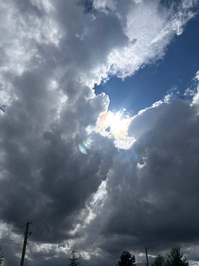 The image depicts a dramatic sky over Pavlovka, Russia, characterized by a turbulent arrangement of clouds. The sun is partially obscured by thick, dark grey clouds, yet its intense light breaks through in a central area, casting bright rays and creating a halo effect with iridescent colors. Two bright, circular lens flares are visible in the lower left quadrant of the image, likely caused by the sun's reflection within the camera lens.
The lower portion of the frame shows the silhouettes of trees and a utility pole with electrical wires stretching across the scene, indicating an inhabited area. The overall atmosphere is one of impending weather change, with the contrast between the dark, heavy clouds and the brilliant, piercing sunlight creating a visually striking and somewhat ominous effect. The specific location within Pavlovka is not discernible from the image itself, but the presence of urban infrastructure suggests it is likely in or near a residential or developed area.