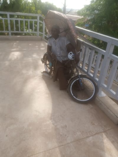 A man sits in a wheelchair on a concrete surface, possibly a walkway or balcony, overlooking a slightly blurred natural background of trees. He is holding a large umbrella open over himself, suggesting it is sunny or potentially raining. The man wears a striped shirt and a cap. His wheelchair appears to be a custom-made motorized tricycle with a rear wheel and two front wheels. The vehicle has a chain drive and a silver exhaust pipe. A white railing runs along the edge of the walkway, with trees and some structures visible beyond it. The lighting suggests it is daytime, possibly late morning or afternoon, with bright sunlight casting shadows. The overall scene conveys a sense of outdoor activity, with the man utilizing his wheelchair for mobility and the umbrella for protection from the elements. No text is clearly visible on any objects in the image.