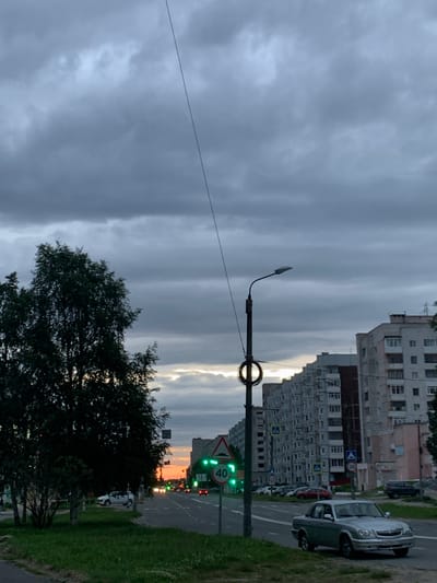 The image shows a street scene in Severodvinsk, Russia, at dusk. The sky is overcast with dark clouds, and a sliver of sunset is visible on the horizon behind apartment buildings. A street lamp with a decorative circular element stands on the left side of the road, and a traffic light displaying green is visible in the distance. A speed limit sign indicating 40 is also present. Several cars are parked along the road and on the sidewalk. A silver sedan is parked on the right side of the road in the foreground. Trees and grass line the left side of the street. The apartment buildings appear to be of Soviet-era construction. No people are clearly visible in the image.
