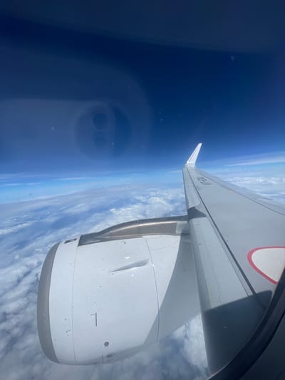 The image shows a view from an airplane window, looking out at the sky and clouds. An airplane wing and engine are visible in the foreground, suggesting the image was taken during flight. The sky is a deep blue, with a layer of white clouds below. There are some smudges and reflections on the window. There is no indication of Neemuch, India, or any urban environment. The image is a generic aerial view from an aircraft.