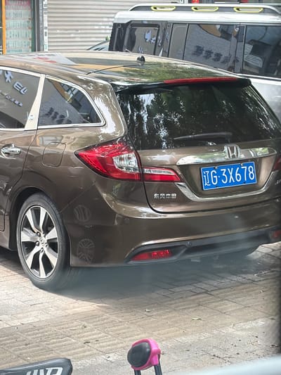 A brown Honda SUV is parked on a tiled street, possibly in Guta, China, judging by the license plate "辽G·3X678". The rear of the vehicle is visible, along with part of its wheel and tire. The side of a white van is also visible to the left of the SUV. The surrounding environment appears to be urban.