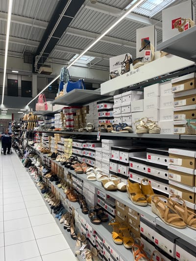 The image shows a shoe store located in Saint-Brice-Courcelles, France at the coordinates (latitude: 49.283, longitude: 4). The store's interior is brightly lit, and the walls are white. There are many shelves filled with shoes. The shelves are made of metal and are arranged in a staggered pattern. The shelves are filled with a variety of shoes, including sandals, sneakers, and boots. Some shoes are in boxes while others are displayed on shelves. There are no people in the store. The image is taken from the right side of the store, looking toward the back. The store is clean and organized. The image shows the variety of shoes for sale, including a woman's shoe on the left side of the image that appears to be on sale for 14 euros. The image is well lit and shows the store's layout. The image shows a variety of shoe styles and colors.