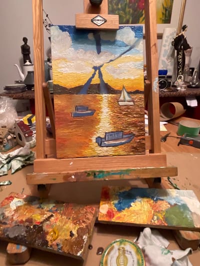 The image shows a painter's studio. An easel holds a painting of sailboats in a harbor scene. Other paintings are visible, as well as art supplies, including paint tubes, brushes, and palettes covered in paint. There is no evidence of the location being Maidstone, United Kingdom.