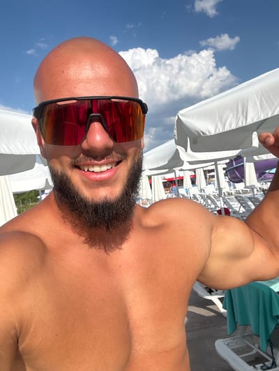 A man with a shaved head and a beard smiles at the camera, wearing red-tinted sunglasses. He is outdoors, with a blue sky and white clouds visible in the background, suggesting a sunny day. Behind him, there are white beach umbrellas and lounge chairs, and a glimpse of a purple water slide. The man is flexing his bicep, and his torso is exposed to the sun. The scene appears to be at a water park or a resort. The location is identified as Ivanyane, Bulgaria.