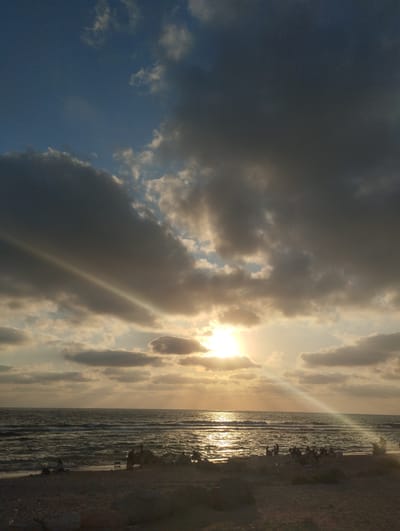 The image depicts a beach scene in the city of Hof HaCarmel Regional Council, Israel, during sunset. The sky is filled with a dramatic mix of dark, heavy clouds and patches of bright blue sky, with the sun's rays piercing through, creating a luminous effect on the water. The sea is moderately wavy, reflecting the golden hues of the setting sun. On the sandy shore, a small group of people are visible, some sitting and others standing, seemingly enjoying the view. The foreground shows a sandy beach with scattered rocks and vegetation. The overall atmosphere is serene and picturesque, characteristic of a coastal sunset.