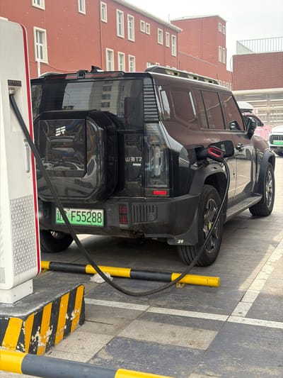 The image shows a black electric vehicle (EV) charging at a public charging station. The location appears to be Guta, China, based on the vehicle's license plate. The EV is parked next to a sidewalk with parking space markings and yellow bollards. The charging cable connects the car to the charging station. Buildings are visible in the background. The scene suggests a common urban setting with infrastructure to support electric vehicle use.