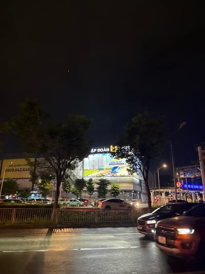 The image depicts a night scene in Thuận An, Vietnam. The perspective is from the sidewalk, looking across a street towards a building and a row of cars. The time of day is clearly night, illuminated by streetlights and the lights from the building and vehicles.
The building in the background is brightly lit, displaying a large advertisement with text in Vietnamese. The advertisement features an image of a modern building complex and includes the phrases "THUẬN AN CƯ" and "100M ĐẾN GA METRO SỐ 2," suggesting it's a real estate development near a metro station. Another sign on the building reads "ẬP ĐOÀN," which translates to "Group" or "Corporation." Near the bottom of the image, a yellow sign with a phone number "0909.642.648" and the text "MỜI QUẢNG CÁO" (Invitation for Advertising) is visible, indicating a location where advertising services are offered.
On the right side of the image, several cars are parked or driving on the street. One car, possibly a dark-colored sedan, is prominent in the foreground, its headlights illuminated. The street itself appears wet, suggesting recent rain or damp conditions, reflecting the surrounding lights.
In the foreground, trees line the sidewalk, partially obscuring the view of the building and street. The overall atmosphere is that of a busy urban environment at night, with the prominent advertising and traffic suggesting commercial activity. There are no discernible people in the image.