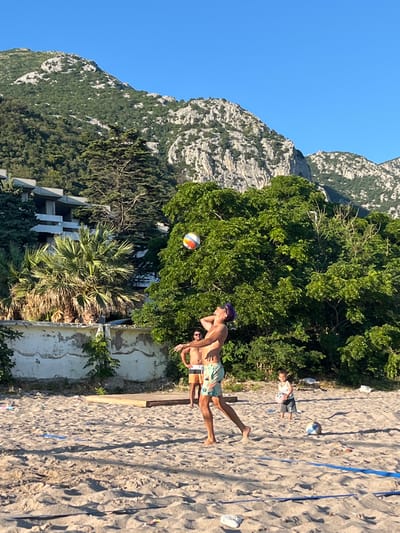 A group of people are playing volleyball on a sandy beach, with a lush green mountain range and a partially constructed building in the background. The sky is clear and blue. In the foreground, a man in swim trunks is in mid-air, spiking a volleyball. Another man stands behind him, and a young child is visible further down the beach. The beach is littered with some debris, and a blue volleyball net is set up. The overall scene suggests a recreational activity in a scenic coastal location.