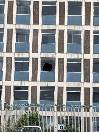 The image shows a close-up, eye-level, full shot of the exterior of a multi-story building in Crewe, United Kingdom. The building features a repeating pattern of windows and textured vertical panels. The windows are divided into upper and lower sections by blue panels. One window on the third floor from the bottom, second column from the right, is broken, with a large, irregular black shape obscuring the glass. Below the windows, a metal fence runs horizontally across the frame, topped with decorative elements that resemble leaves. Beyond the fence, greenery and a white vehicle are visible. A CCTV camera is mounted on the building to the left of the fence. A sign for "KENT HOUSE" is visible on the building in the upper right quadrant.