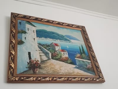 The image shows a framed painting of a coastal village scene, likely in a Mediterranean setting. The painting depicts white-washed buildings with red tile roofs perched on a hillside overlooking a bright blue sea. Lush green foliage and a winding path with stairs are visible, adding depth and texture to the scene. A vase of colorful flowers sits on a ledge in the foreground. The painting is encased in an ornate gold frame. The background is a plain white wall with a subtle architectural molding near the top. The painting is hung at an angle, suggesting it is not perfectly level or is viewed from a side perspective. The location of the painting in the city of Ayagöz, Kazakhstan, is not evident from the visual information provided in the image.
