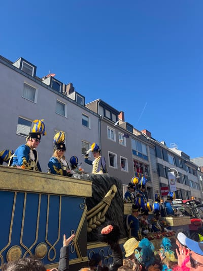 The image shows a festive parade in Köln, Germany, at the coordinates 50.926, 6.958. The scene is captured from a low angle, showcasing a procession of people in colorful costumes riding on a decorated float. The float features a blue and gold design, adorned with ornate rope details. Several people in blue and yellow costumes are visible, with one woman seated on the float, looking towards the viewer. Other participants are visible in the distance, walking along a street lined with buildings. The sky is clear and blue. The buildings are multi-story and appear to be residential, with windows visible. The atmosphere is joyous and celebratory, suggesting a carnival or parade event.  The image captures a moment of festivity and celebration in the city.