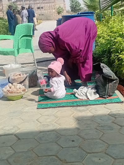 An outdoor scene in Kwazailewa, Nigeria, depicts a woman and a child on hexagonal paved ground. In the foreground, a young child, dressed in a white patterned long-sleeved garment and a pink knitted hat with a pom-pom, sits on a green and orange woven mat. The child holds a clear plastic bottle with a red label in its left hand and looks towards the camera. To the child's right, a crumpled white cloth and a dark fabric bag are present on the mat. A woman wearing a deep purple hijab and a matching long garment is bent over the mat, facing left, with her back partially towards the camera, appearing to attend to the child or items on the mat.
To the left of the mat, a partially visible light green plastic chair stands near several containers: two clear plastic buckets containing various items (one with white, fluffy material, another with brownish, irregularly shaped contents), and a light green bowl holding numerous spherical, light-colored food items. In the mid-ground, behind the woman, two large blue plastic barrels are visible. Further in the background, a group of approximately five to six men, dressed in various traditional attire, stand conversing on a paved path. Behind them, a large, multi-story building under construction, primarily concrete framing, is visible. The scene is illuminated by strong sunlight, casting distinct shadows. Green bushes and plants are present on the right and left sides of the image, with palm fronds visible at the top right.