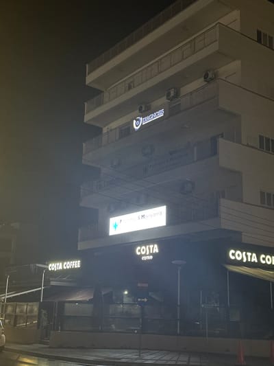 The image depicts an urban street scene at night in Egkomi Lefkosias, Cyprus, focusing on a multi-story commercial building. The sky is dark, indicating nighttime, and the scene is illuminated by artificial lights from streetlights and business signs.

On the ground floor of the building, a prominent Costa Coffee store is visible with multiple brightly lit signs reading "COSTA COFFEE." A large horizontal sign spans the facade, and a smaller vertical sign also displays the brand. An awning extends over the entrance, and the interior, though dimly lit, shows some seating through its glass windows.

Adjacent to Costa Coffee, an illuminated sign advertises medical practices: "Panayiotis & Maroyiannis," "Pneumonologist & Melegiannis," and "General Practitioner," accompanied by a blue star logo. Higher up on the building's facade, an illuminated sign clearly displays the word "DIAGNOSIS" with a circular logo. A partially visible sign with the text "Loumen" can also be seen further up the building.

The building itself features balconies with railings and several external air conditioning units. The foreground shows a dark street or sidewalk area with a visible lamppost. An arrow sign pointing to the right is located near the Costa Coffee entrance. The scene appears quiet and devoid of active human presence.