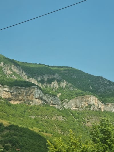 The image shows a mountainous landscape with rocky cliffs and lush green vegetation under a clear blue sky. A power line stretches diagonally across the upper part of the image. The terrain is characterized by steep slopes, layered rock formations, and dense forests. The lighting suggests it is daytime, with some areas of the mountain in shadow, indicating the sun's position. The scene is natural and undeveloped, with no visible signs of urban structures except for a very small portion of a reddish-brown roof peeking out from the bottom left corner, possibly part of a distant building or dwelling. The overall impression is one of rugged natural beauty. This image is likely captured in or near Ochindol, Bulgaria, given the geographical context provided.
