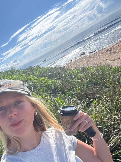 A female individual is captured in a selfie-style photograph, positioned outdoors in a coastal environment within Wollongong City Council, Australia. She has blonde hair, light eyes, and is wearing a grey t-shirt and a faded grey baseball cap with "X CUB" embroidered in orange/tan lettering. A silver hoop earring is visible on her left ear, a silver band ring on her left ring finger, and a silver bracelet on her left wrist. She holds a brown patterned cardboard coffee cup with a black plastic lid in her left hand, displaying a logo featuring a tree and the word "FOSTERS". The immediate foreground shows dense green coastal vegetation, including grasses and shrubs. Beyond this vegetation, a sandy beach transitions to the ocean, where small waves break under bright sunlight, creating glints on the water's surface. A small, dark-colored boat with at least two occupants is visible in the water. The sky is bright blue with scattered white, wispy clouds. The scene is illuminated by natural sunlight.