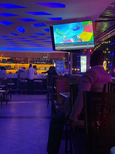 The media file depicts a dimly lit bar or lounge environment, likely in Kimorori, Kenya, during the evening or night. The scene features several patrons and staff amidst a vibrant play of artificial lights, predominantly blue and yellow.
Subjects include various people seated at tables and a long bar counter. One individual in a light-colored suit jacket is prominently seated at a table in the foreground, facing away from the camera. Several other people are visible seated on stools at the bar counter, with a bartender or staff member behind it. The bar is well-stocked with numerous bottles of alcohol, illuminated by yellow backlighting.
A large flat-screen television is mounted on a wall to the left, displaying colorful animated content resembling a game board with objects like boats and dice. The TV screen also shows an interface with text such as "Exploring", "327 SOUNDCITY", "Viewers Instant Playlist", "Tee Choice", and "BTS". Time indications like "15:00", "20:00", and "21:40" are present, along with the date "15:07 Tue 17 Jan". Another visible phrase on the screen is "Press HELP for Mail".
The room features distinctive decor, including a wall with intricate blue geometric patterns that are backlit, casting a soft glow. Overhead, string lights with small bulbs add to the festive atmosphere. A sign behind the bar partially reads "ENJOY & COLD". The overall activity suggests a social gathering, with people relaxing, possibly watching content on the screen, and enjoying drinks at the bar in a casual setting.