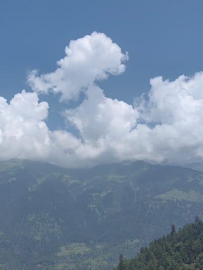 The image displays a panoramic view of the Solang Valley in India, characterized by lush green mountainous terrain under a bright blue sky dotted with fluffy white clouds. The foreground features a dense forest of pine trees on a steep slope. In the mid-ground, rolling hills covered in a mixture of evergreen trees and grassy meadows descend into the valley. Some areas of cultivation are visible on the gentler slopes. The background is dominated by layered mountain ranges, their peaks partially obscured by wispy clouds. The overall scene evokes a sense of natural beauty and tranquility, typical of a mountainous landscape. There are no people or man-made structures clearly visible in this particular shot, focusing solely on the natural environment.