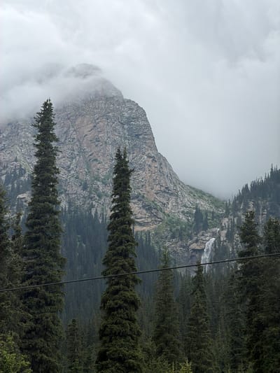 The image shows a mountainous landscape in Kyrgyzstan. In the foreground, several tall, dark green coniferous trees are visible, some partially obscuring the view. A thin black cable runs horizontally across the mid-ground. Behind the trees, a large, rugged mountain with rocky faces and patches of green vegetation rises into a cloudy sky. A waterfall cascades down the side of the mountain in the background. The overall atmosphere is overcast and damp, suggesting a cool climate.