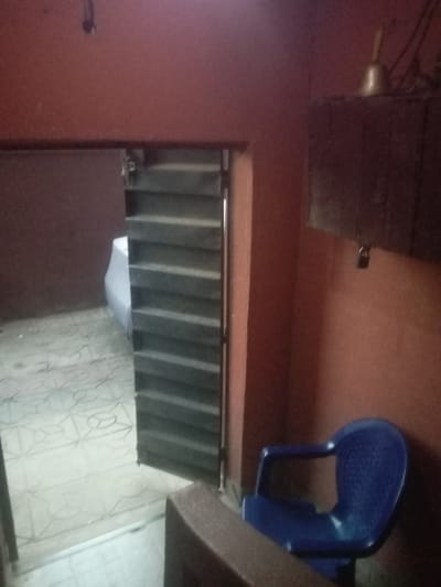 This image captures an interior scene, likely a room or a transitional space within a building in Iyiowa Odekpa, Nigeria. The walls are painted a reddish-brown hue.
In the foreground to the right, a bright blue plastic chair is visible, angled towards the corner of the room. Above the chair and mounted on the wall is a dark, box-like structure, possibly a cabinet or shelf, from which a padlock is hanging. Perched on top of this structure is a brass-colored bell.
To the left, a doorway is present, partially obscured by what appears to be a slatted or louvred door or screen, possibly made of wood or metal. Through this opening, a glimpse of another space is visible, showing a patterned tiled floor similar to the one in the foreground. Part of a large white object, possibly a vehicle or a piece of furniture, can be seen beyond the doorway.
The tiled floor in the main space features a light-colored pattern with geometric or abstract shapes. In the upper left of the image, some dark wiring or a fixture hangs from the ceiling. The lighting in the scene is dim, suggesting an indoor setting without strong natural light. There is no discernible activity taking place, and no visible text.